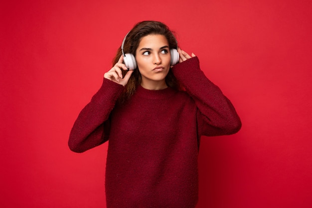 Beautiful young brunette curly woman wearing dark red sweater isolated over red background wall wearing white bluetooth headphones listening to music and having fun looking up to the side and thinking