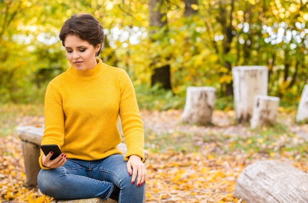 Beautiful young brunette girl posing in the autumn park with a mobile phone in her hands