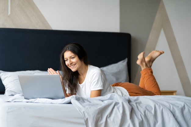 Beautiful young brunette girl with a laptop lying on the bed. Stylish modern interior. A cozy workplace. Shopping on the Internet.