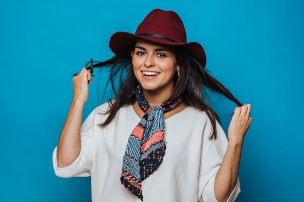 Beautiful young brunette holds her hair, looking happy, dressed in white sweater, burgundy hat with a brim and coloured neckerchief on the neck, smiles broadly. isolated over blue background