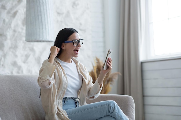 Beautiful young brunette woman wearing glasses at home in living room shocked received good news