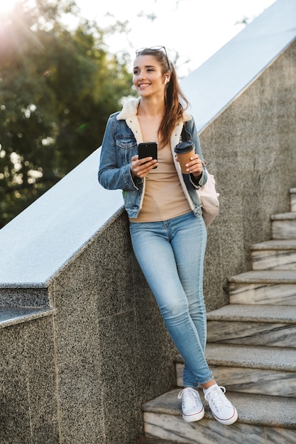 Beautiful young brunette woman wearing jacket, carrying backpack walking outdoors, using mobile phone, holding coffee cup