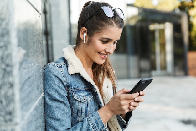 Beautiful young brunette woman wearing jacket, carrying backpack walking outdoors, using mobile phone