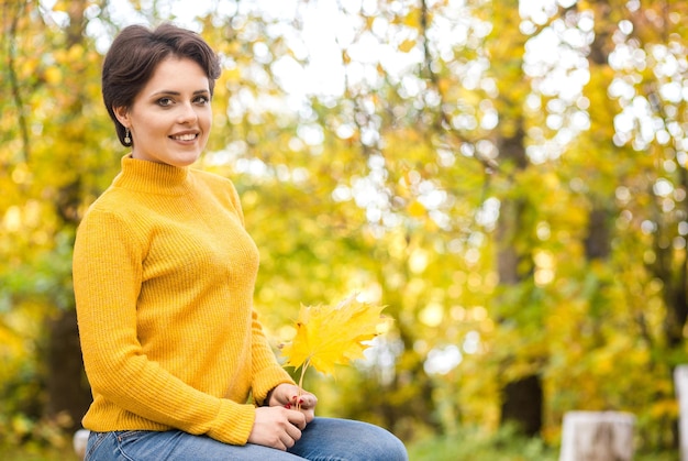 Beautiful young brunette woman in a yellow knitted sweater posing in an autumn park with a bouquet of leaves