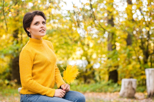 Beautiful young brunette woman in a yellow knitted sweater posing in an autumn park with a bouquet of leaves