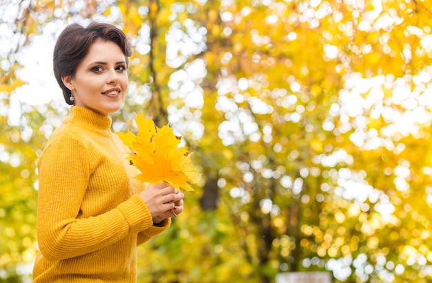 Beautiful young brunette woman in a yellow knitted sweater posing in an autumn park with a bouquet of leaves