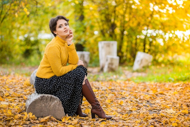 Beautiful young brunette woman in a yellow knitted sweater skirt and boots posing in the autumn park and smiling