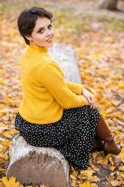 Beautiful young brunette woman in a yellow knitted sweater skirt and boots posing in the autumn park