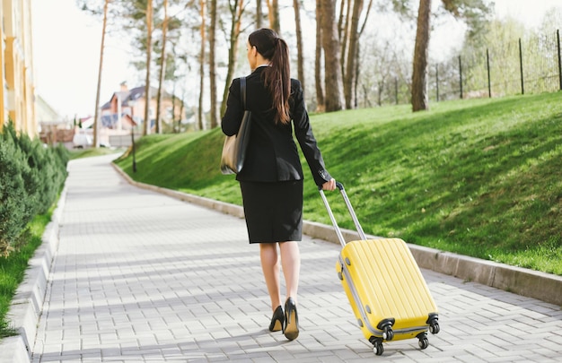 Beautiful young business woman in a suit and in shoes walking on a city street with a yellow travel suitcase