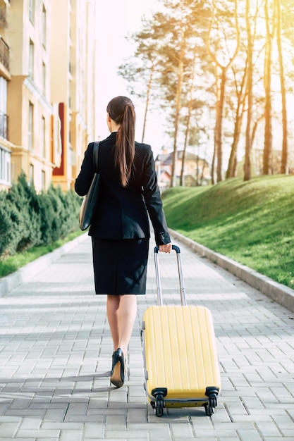 Beautiful young business woman in a suit and in shoes walking on a city street with a yellow travel suitcase