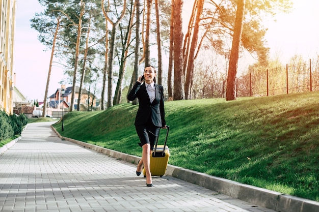 Beautiful young business woman in a suit and in shoes walking on a city street with a yellow travel suitcase