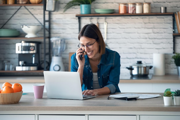 Beautiful young business woman talking with her mobile phone while working with computer in the kitchen at home