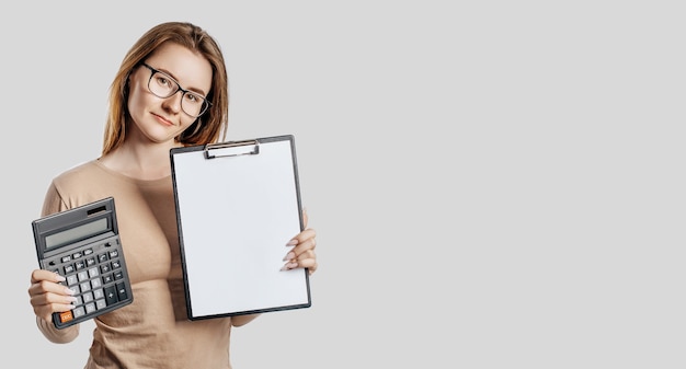 Beautiful young business woman wearing glasses holds calculator and clipboard with mock up blank space isolated on gray space