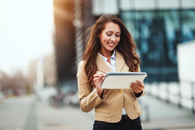 Beautiful young businesswoman is holding tablet in her hands against urban city background.