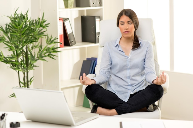 Beautiful young businesswoman practicing yoga in the office. She sits on lotus pose in a chair in front of her desk.