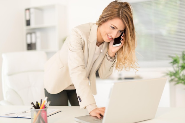 Beautiful young businesswoman sitting in the office, using phone and writing on laptop.