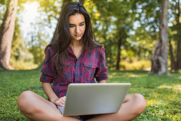 Beautiful young Caucasian happy woman wearing plaid shirt sitting on grass in front of laptop pc working on her laptop computer shopping and wireless communication Freelance female working outdoor