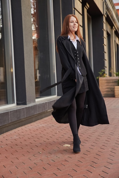 Beautiful young caucasian red headed girl in stylish black coat walking outdoors