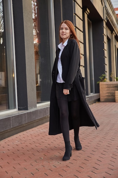 Beautiful young caucasian red headed girl in stylish black coat walking outdoors