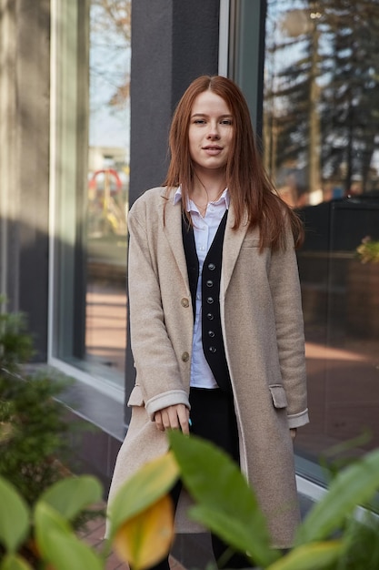 Beautiful young caucasian red headed woman in beige coat with long hair standing outdoors behind green plants. attractive female street portrait at fall. autumn lifestyle, stylish fashion outfit