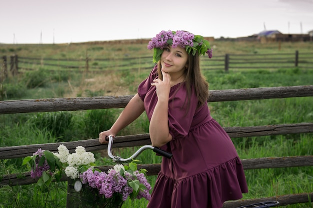 A beautiful young Caucasian smiling happy girl in purple dress in a wreath and with a bicycle a basket of blooming lilac in the countryside. Model plus size. Spring, rustic style. Circlet of flowers