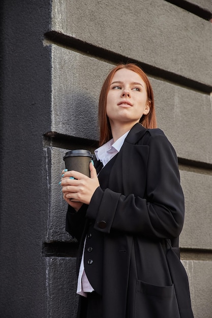 Beautiful young caucasian woman in stylish beige coat drinking coffee outdoors