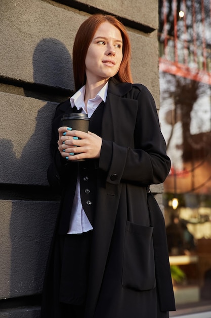 Beautiful young caucasian woman in stylish beige coat drinking coffee outdoors