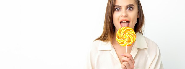 Beautiful young caucasian woman wearing a white shirt licking a lollipop on a white background