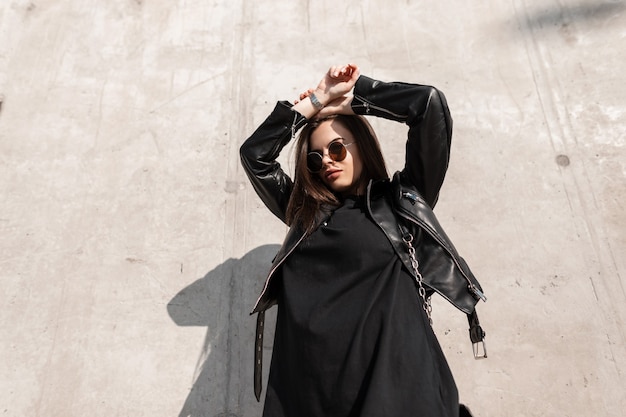 Beautiful young cool caucasian girl model with fashionable hipster sunglasses in stylish black clothes with a leather jacket and sweatshirt on a concrete wall background in sunlight