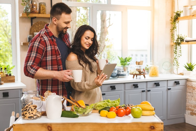 Beautiful young couple is talking,  smiling while eating tea or coffee and drinking in kitchen at home. Using tablet.