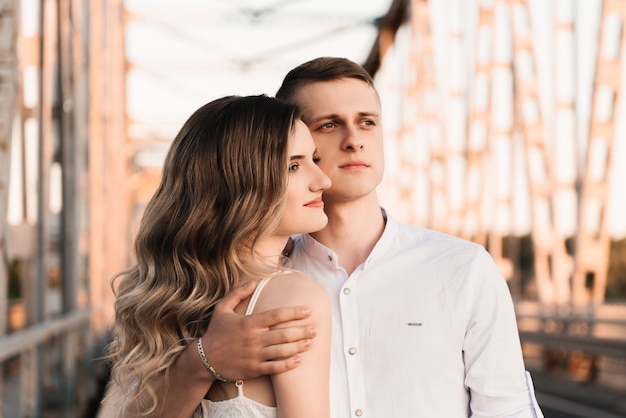 A beautiful young couple in love, a man and a woman, embrace, kiss on a large metal bridge at sunset.