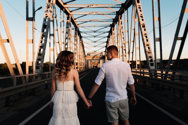 A beautiful young couple in love a man and a woman embrace kiss on a large metal bridge at sunset