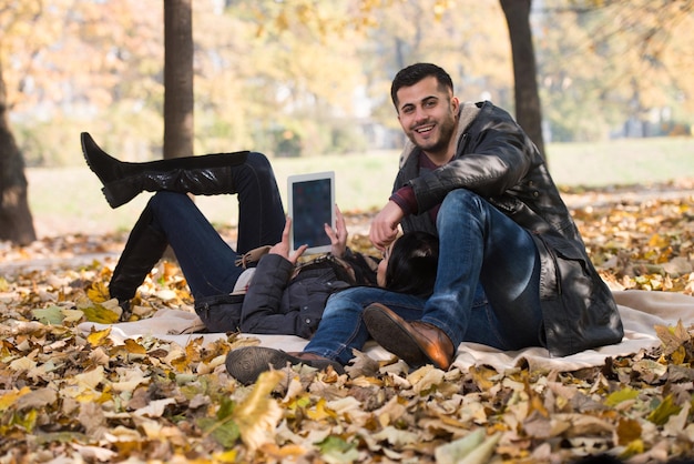 Beautiful Young Couple Sitting In The Park On A Beautiful Autumn Day  They Are Using Internet Via Digital Tablet