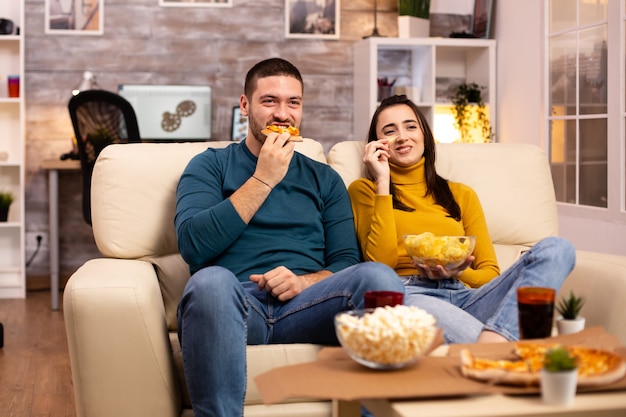 Beautiful young couple watching TV and eating fast food takeaway in the living room