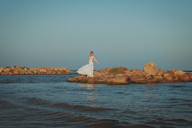 Beautiful young elegant pregnant blond woman in a blue hat walks on the beach, seashore at sunset