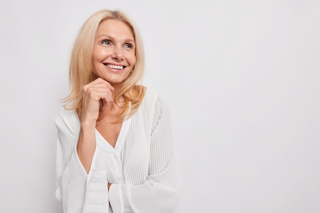 Beautiful young European woman of middle age smiles gently keeps hand under chin looks away with dreamy expression wears silk blouse isolated over white wall copy space for advertisement