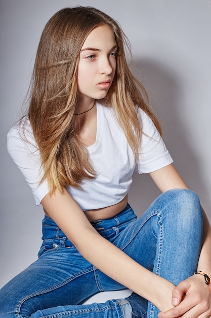 Beautiful young fashion model girl in blue jeans sitting on the floor on a white background. Natural beauty girls, teen and adolescent issues.