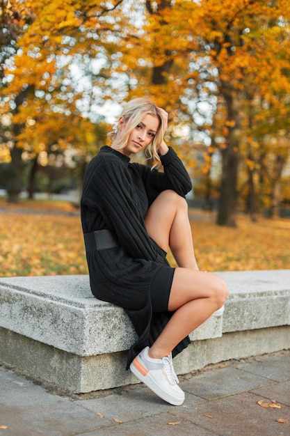 Beautiful young fashionable fresh autumn girl in a fashion black knitted dress with white sneakers sits in an autumn city park with yellow foliage