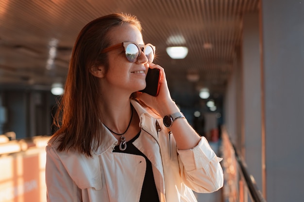 Beautiful young fashionable woman in sunglasses with a mobile phone smartphone in her hands communicates on the phone on the urban urban background of the tunnel in the sunset rays.