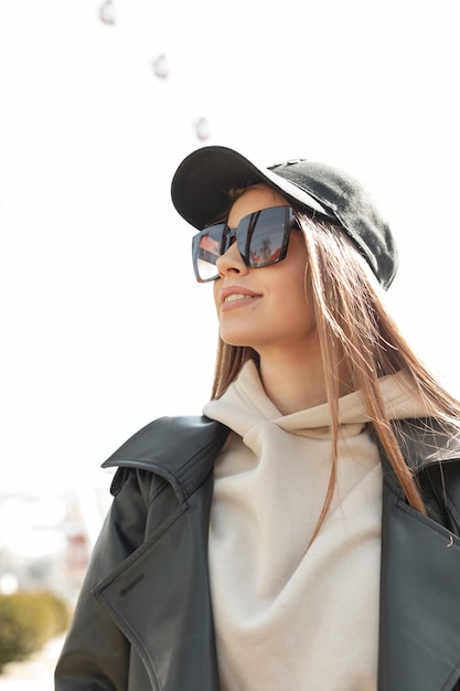 Beautiful young fashionable woman with cool stylish sunglasses in a fashion black leather jacket hoodie and trendy black cap walks on a sunny warm spring day