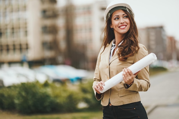 Beautiful young female engineer with white helmet and blueprints in her hands in front of office district.