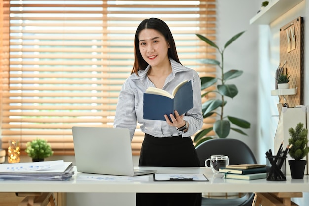 Beautiful young female entrepreneur holding diary planner standing at her working desk and using laptop computer