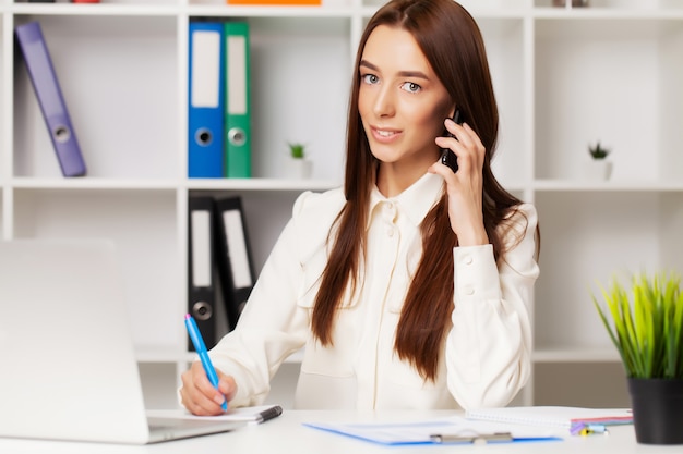 Beautiful young female manager talking on the phone while working on laptop in the office