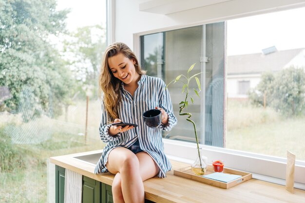 beautiful young female siting on kitchen counter and browsing on mobile phone i modern kitchen