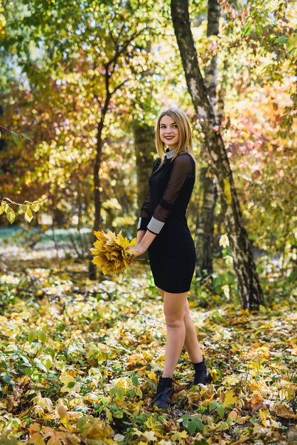 Beautiful young female student in black dress holding a bouquet of leaves in autumn park.