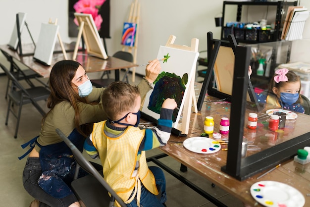 Beautiful young female teacher is helping a little boy to paint on a canvas during art school