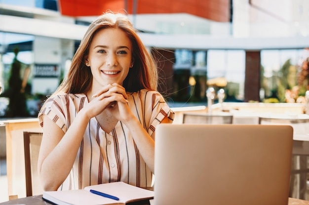 Beautiful young female vlogger doing a vlog outside in a coffee shop with a laptop and a notebook on the table looking at camera smiling.