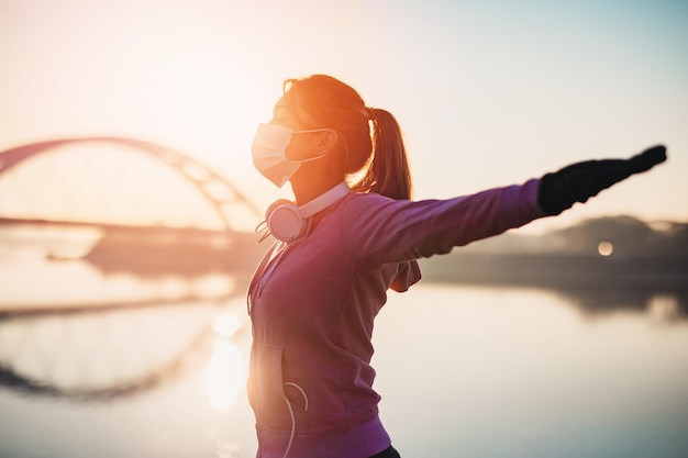 Beautiful young and fit woman in good shape running and jogging alone on city bridge street. She wearing protective face mask to protect herself from virus or allergy infection. Sunset in background.