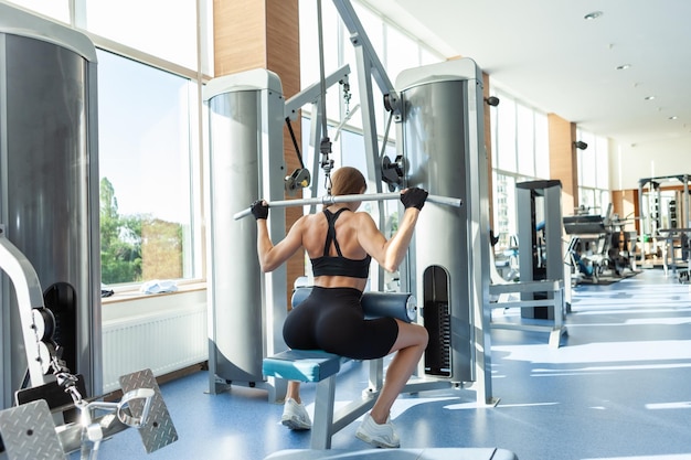 Beautiful young fit woman working out on a pull down lat machine at the gym. Back view