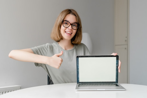 Beautiful Young Freelancer Woman showing Laptop Computer mockup screen and thumb up sitting at home. Freelance Work, online education Concept